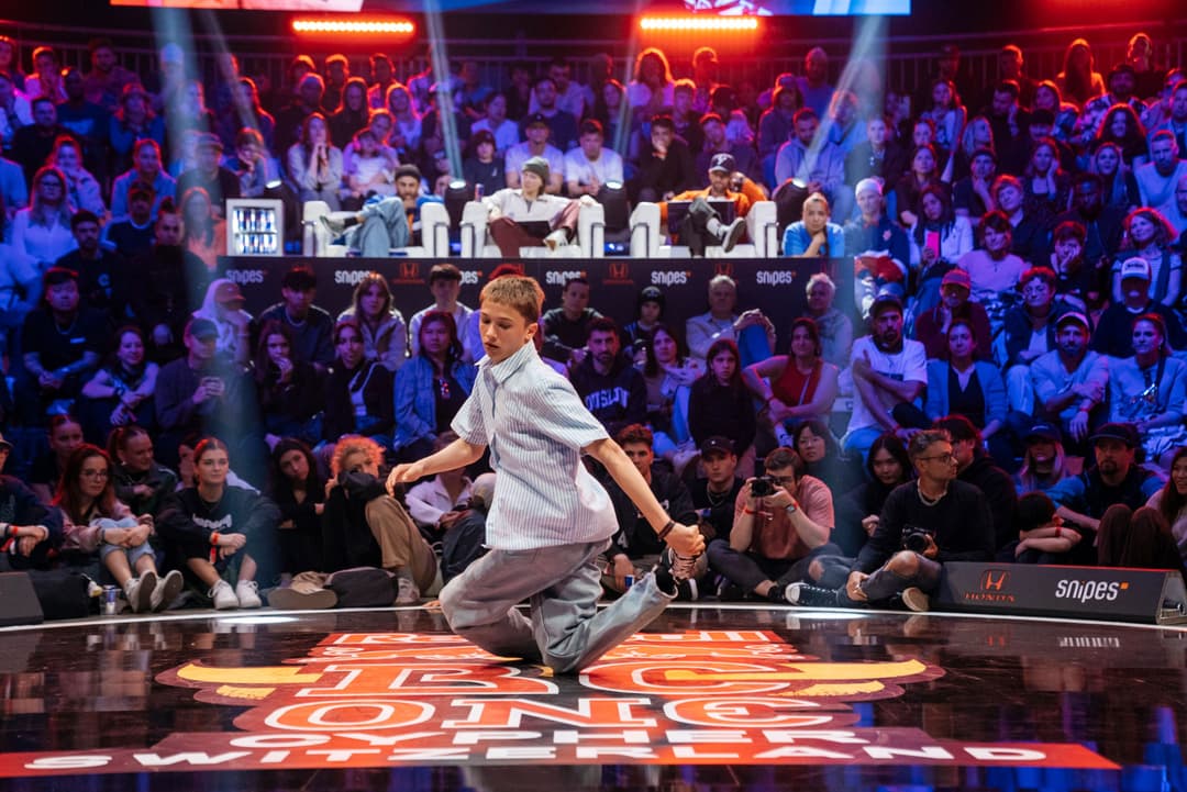 Danseur de breaking en mouvement sur un sol coloré, entouré de spectateurs et d'un jury assis derrière, lors d'une compétition Red Bull BC One