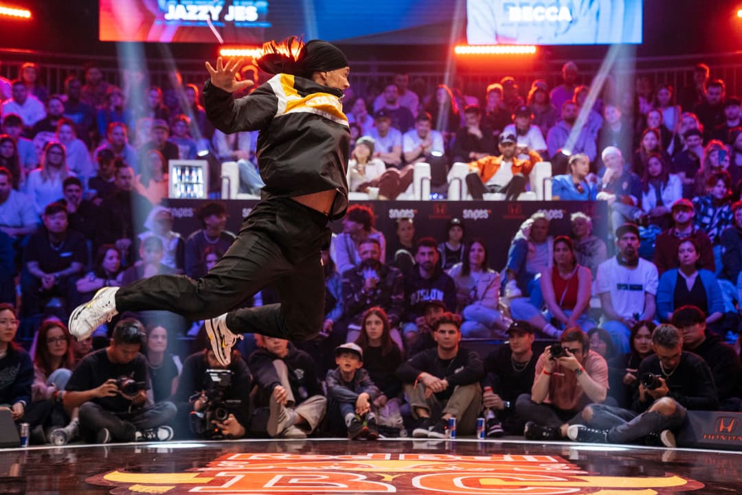 B-boy en costume noir et jaune effectue un saut acrobatique sur la piste de danse du Red Bull BC One à Lausanne, devant un public attentif