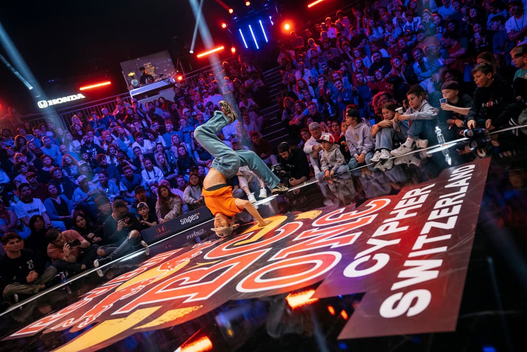 Danseur de breaking en compétition sur scène, effectuant un mouvement acrobatique devant un public nombreux illuminé en bleu et rouge