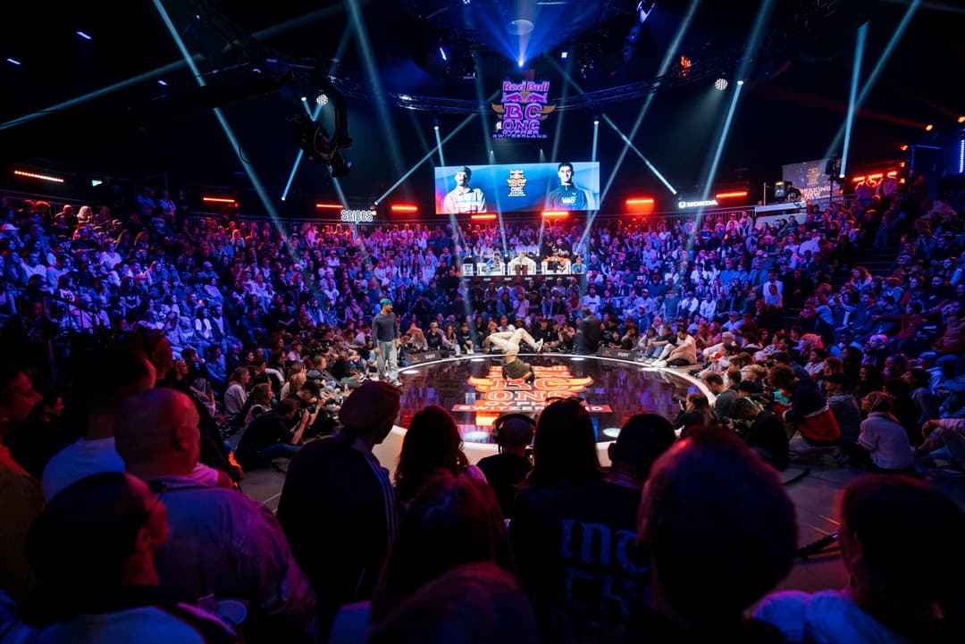 Scène du Red Bull BC One avec une piste circulaire illuminée au centre, entourée d'une foule de spectateurs dans une arène aux lumières bleues et rouges
