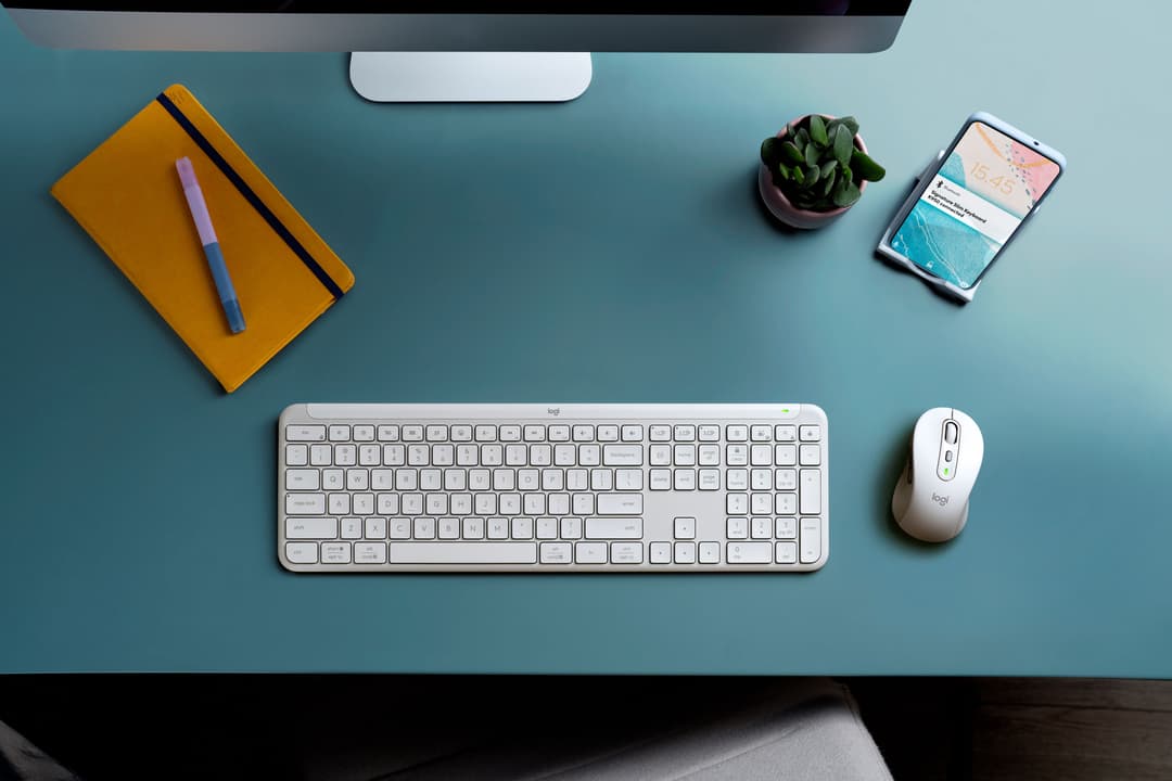 Clavier Logitech Signature blanc avec souris assortie sur un bureau bleu, accompagnés d'un carnet jaune et d'un smartphone