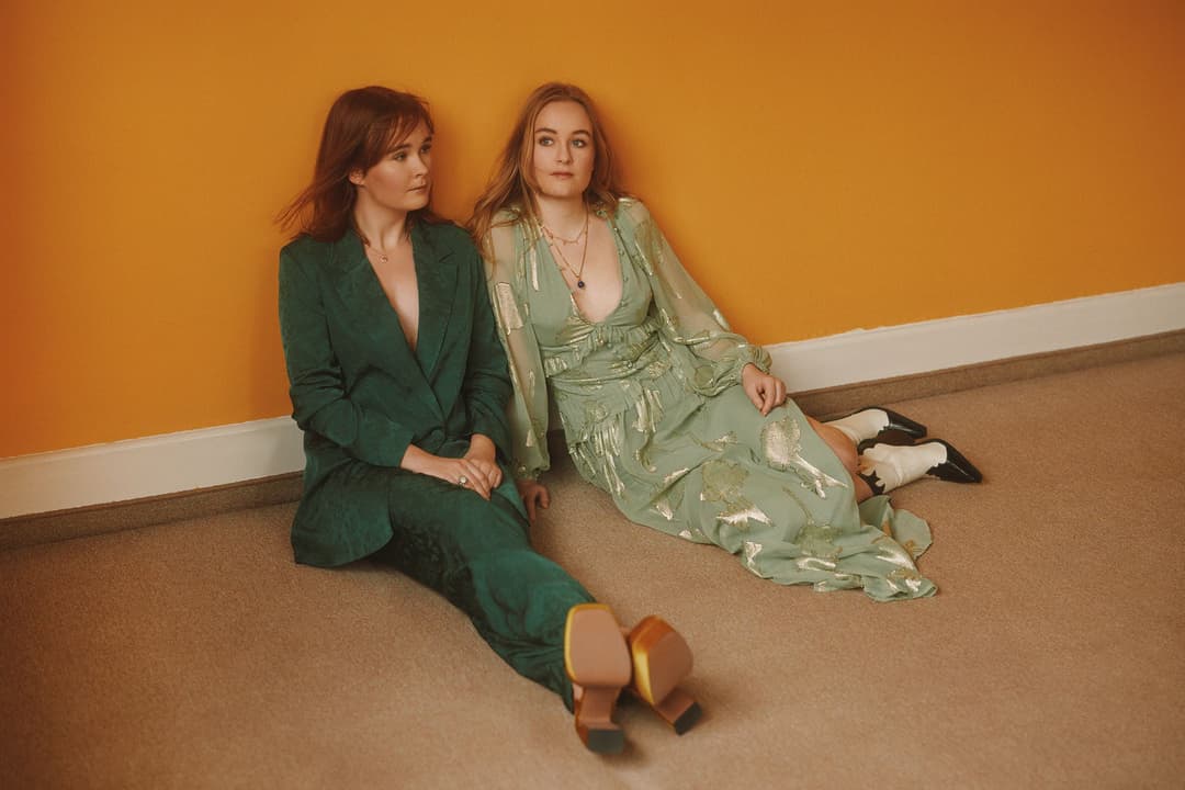 Hannah-Leah et Meghan du duo The Woodgies assis sur le sol devant un mur bicolore orange et blanc