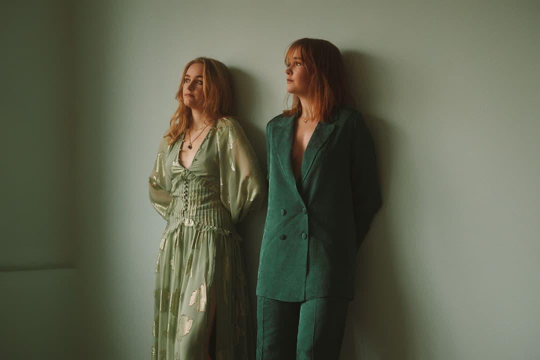 Meghan et Hannah-Leah, les deux membres du duo musical The Woodgies, posent côte à côte devant un mur vert pâle