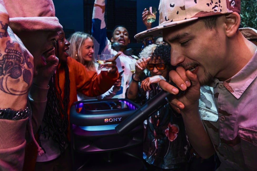 Homme en casquette tenant un microphone lors d'une fête, entouré de personnes, avec un haut-parleur Sony ULT aux éclairages bleus et roses