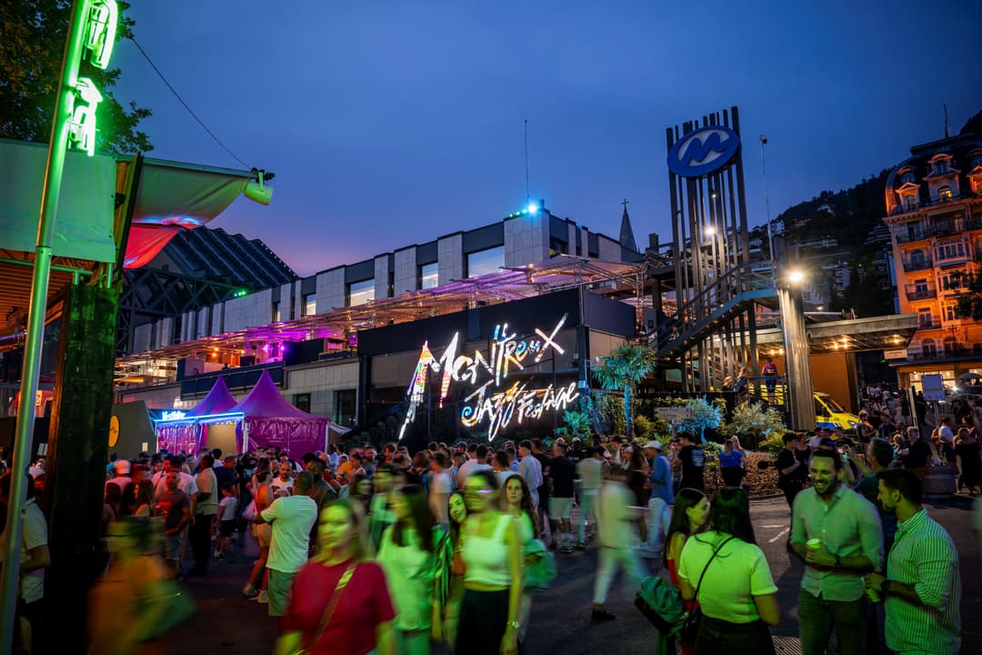 La place du Montreux Jazz Festival de nuit, avec foule colorée illuminée en vert fluo et rose, bâtiments modernes et logo du festival