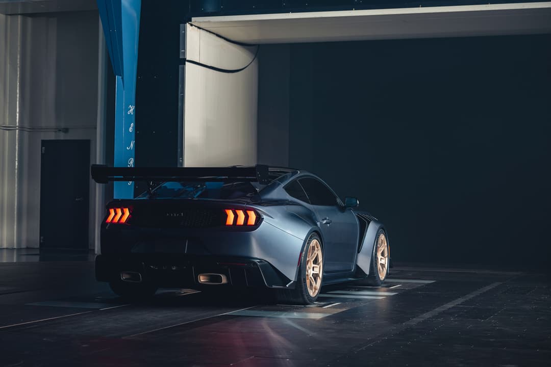 Ford Mustang gris métallisé vue de trois-quarts arrière avec aileron carbone et feux arrière orange allumés, garée en intérieur sous éclairage bleu