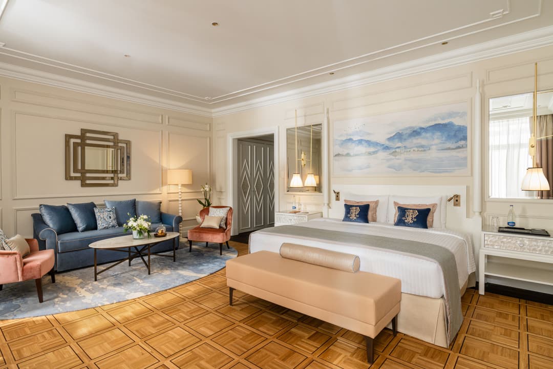 Suite rénovée du Fairmont Le Montreux Palace avec lit blanc, canapé bleu, fauteuils rose et beige, tableau des Alpes au mur