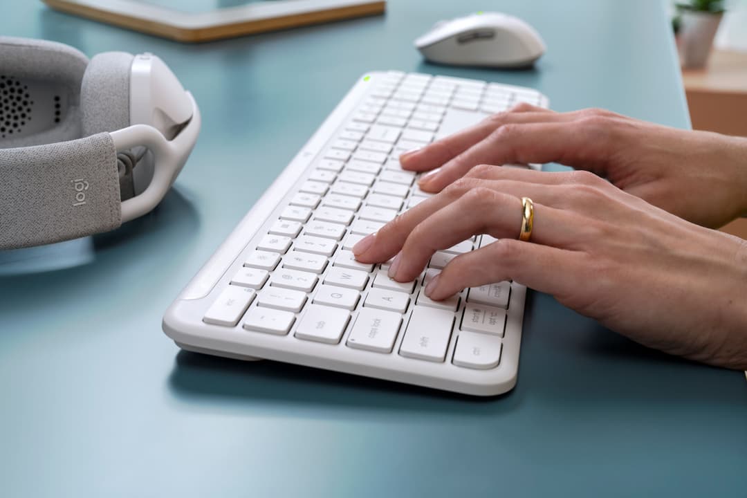 Clavier Logitech blanc avec touches plates, mains posées dessus, environnement bureau minimaliste bleu