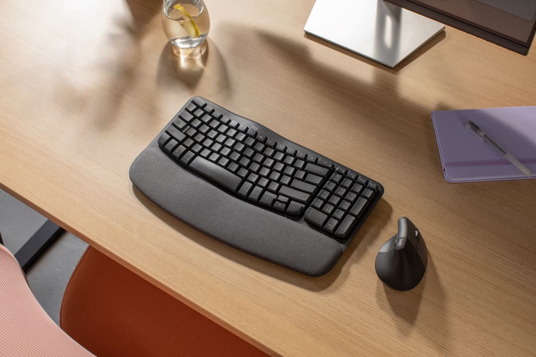 Clavier Logitech Wave Keys gris avec repose-poignets et souris noire sur un bureau en bois, entouré d'écrans et d'accessoires