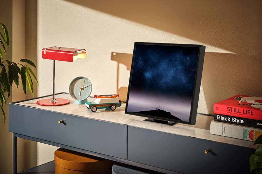 Samsung Music Frame affichant un paysage étoilé, posé sur un bureau gris à côté d'une lampe rouge, d'une horloge et de livres