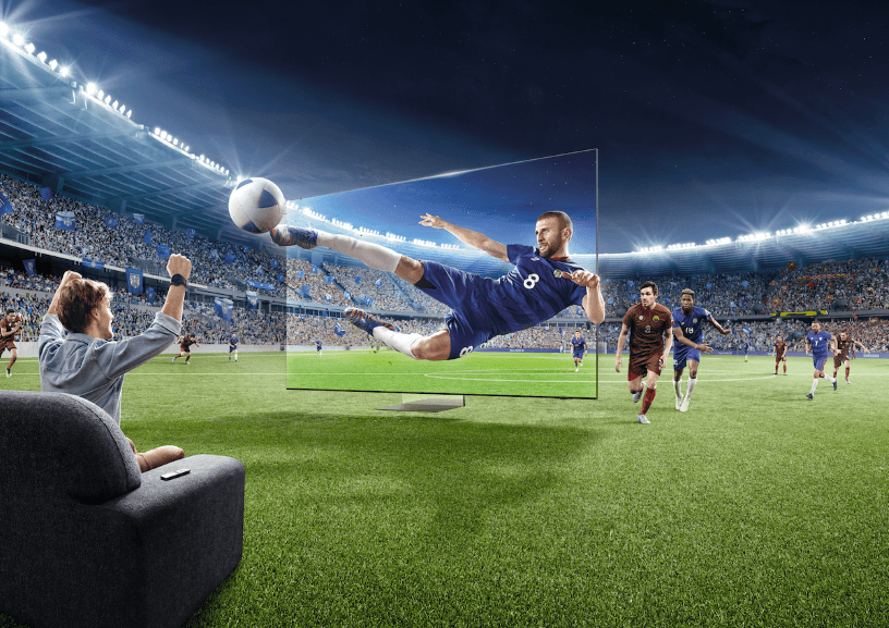 Spectateur assis sur un canapé regardant un match de football sur écran géant Samsung, un joueur en maillot bleu effectuant un tir acrobatique au stade illuminé