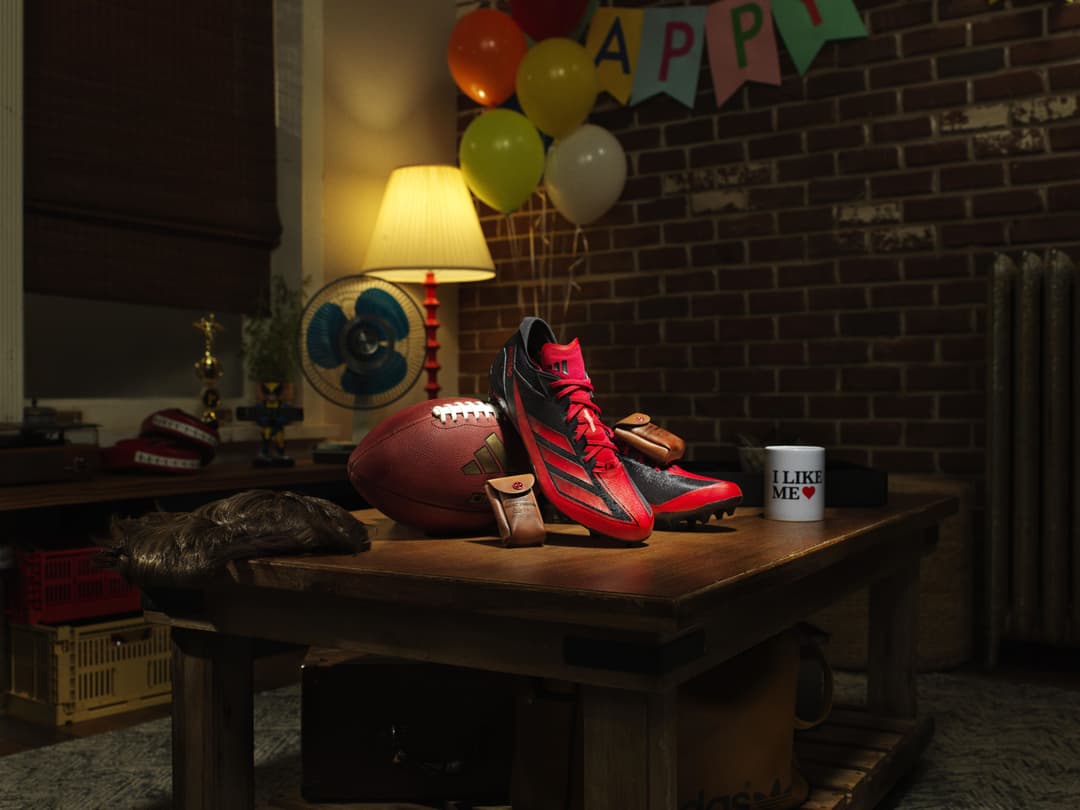 Chaussure de football adidas rouge et noire thématisée Deadpool exposée sur un bureau en bois, aux côtés d'un ballon de football et d'accessoires assortis