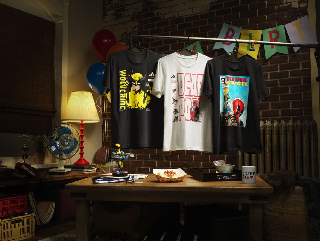 Trois t-shirts Adidas inspirés par Deadpool et Wolverine affichés sur des cintres, avec motifs jaunes et rouges, dans un décor de chambre aux briques apparentes