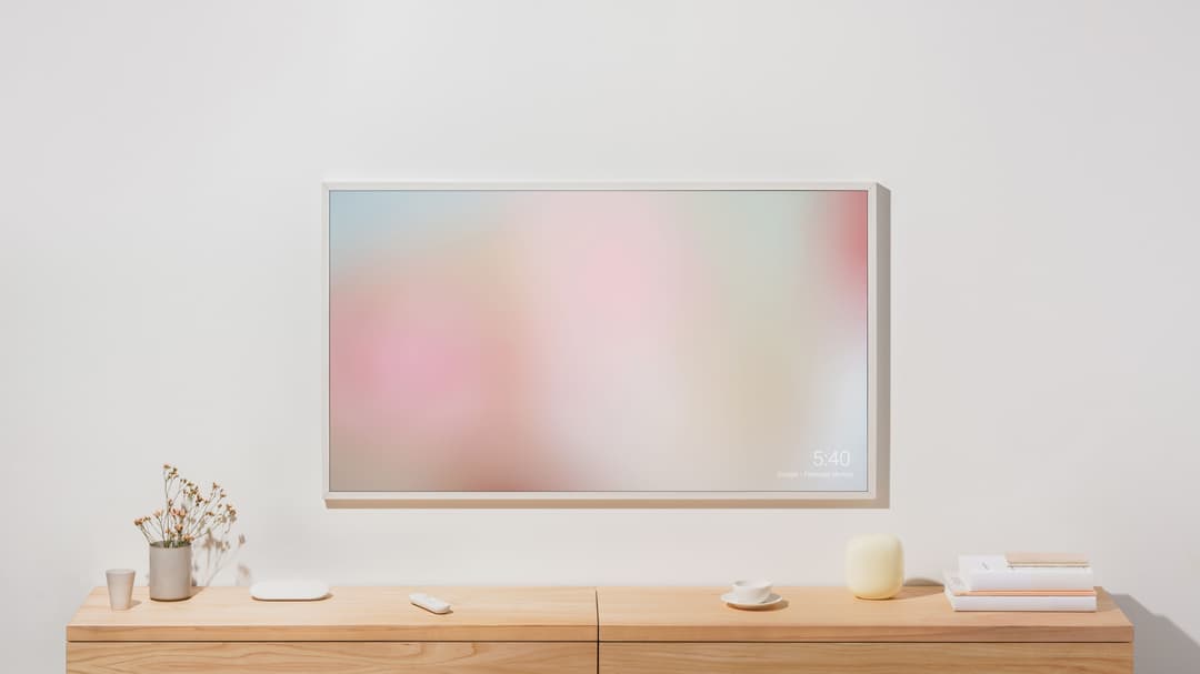 Google TV Streamer blanc posé sur un meuble en bois devant un téléviseur mural affichant un dégradé pastel