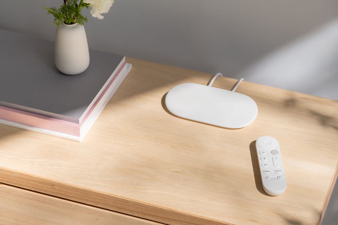 Google TV Streamer blanc avec sa télécommande sur un bureau en bois, à côté d'une souris sans fil et d'un vase de fleurs
