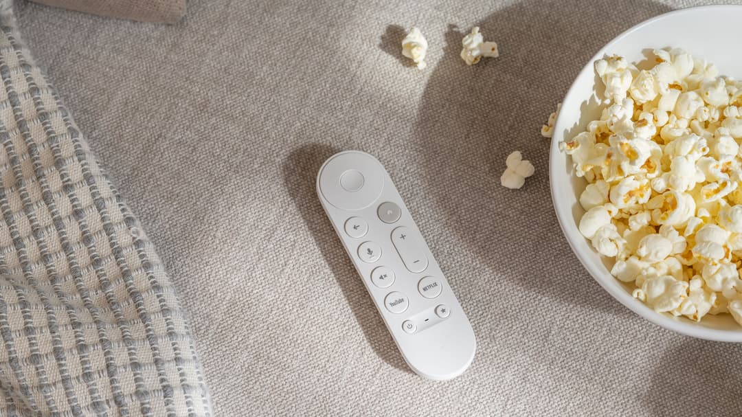 Télécommande Google TV Streamer blanche entourée de pop-corn et d'une couverture, sur fond beige