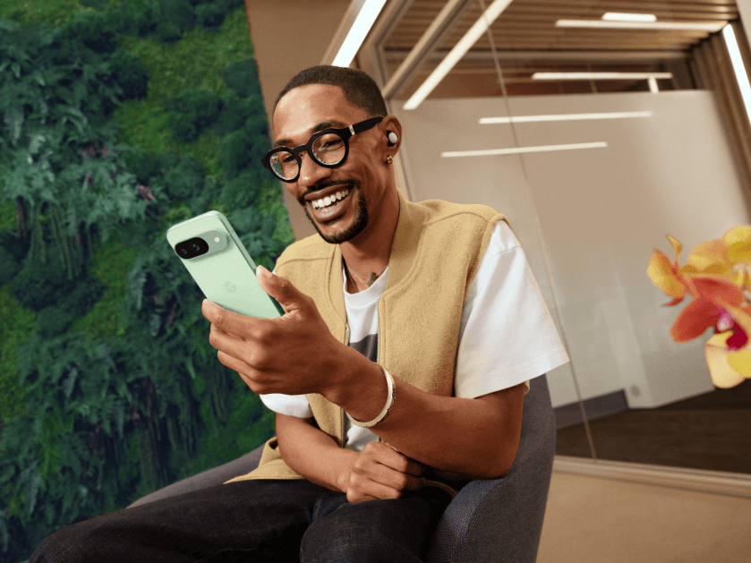 Homme souriant tenant un smartphone Pixel 9 de couleur verte clair, dans un intérieur lumineux avec plante verte en arrière-plan