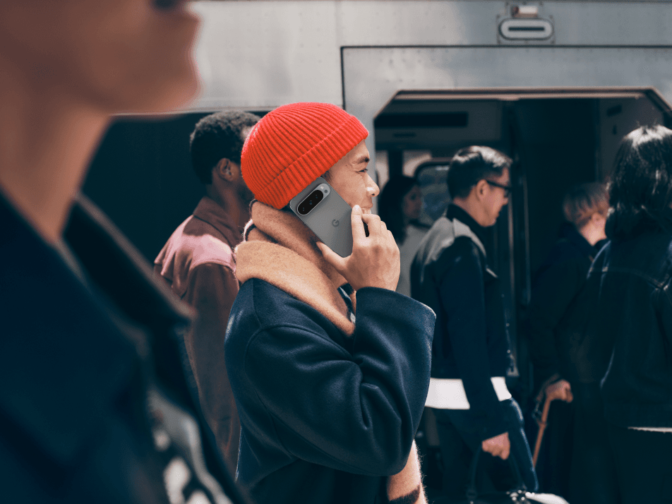 Homme portant un bonnet rouge tenant un smartphone Pixel 9 à l'oreille, entouré d'autres personnes devant un bus
