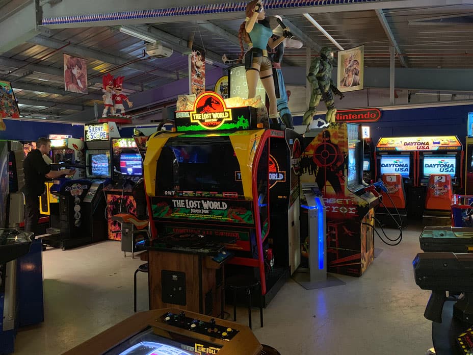 Salle de jeux vidéo rétro avec bornes d'arcade colorées, dont une centrale en jaune et rouge représentant le jeu The Lost World avec une figurine de dinosaure