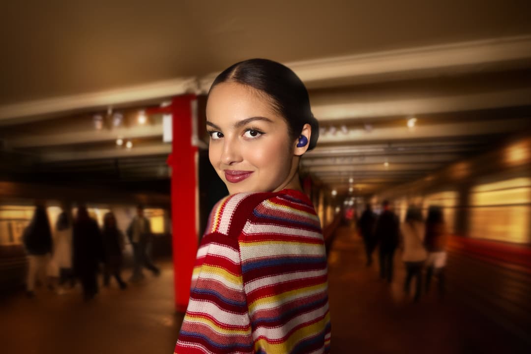 Femme portant des écouteurs Sony LinkBuds de couleur violette, vêtue d'un pull à rayures multicolores, dans un intérieur moderne