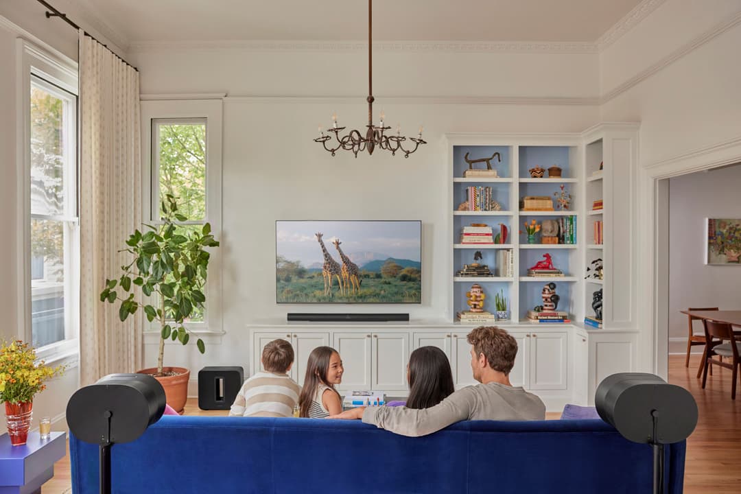 Sonos Arc Ultra montée sous un téléviseur dans un salon blanc avec canapé bleu, quatre personnes assises de dos regardant l'écran