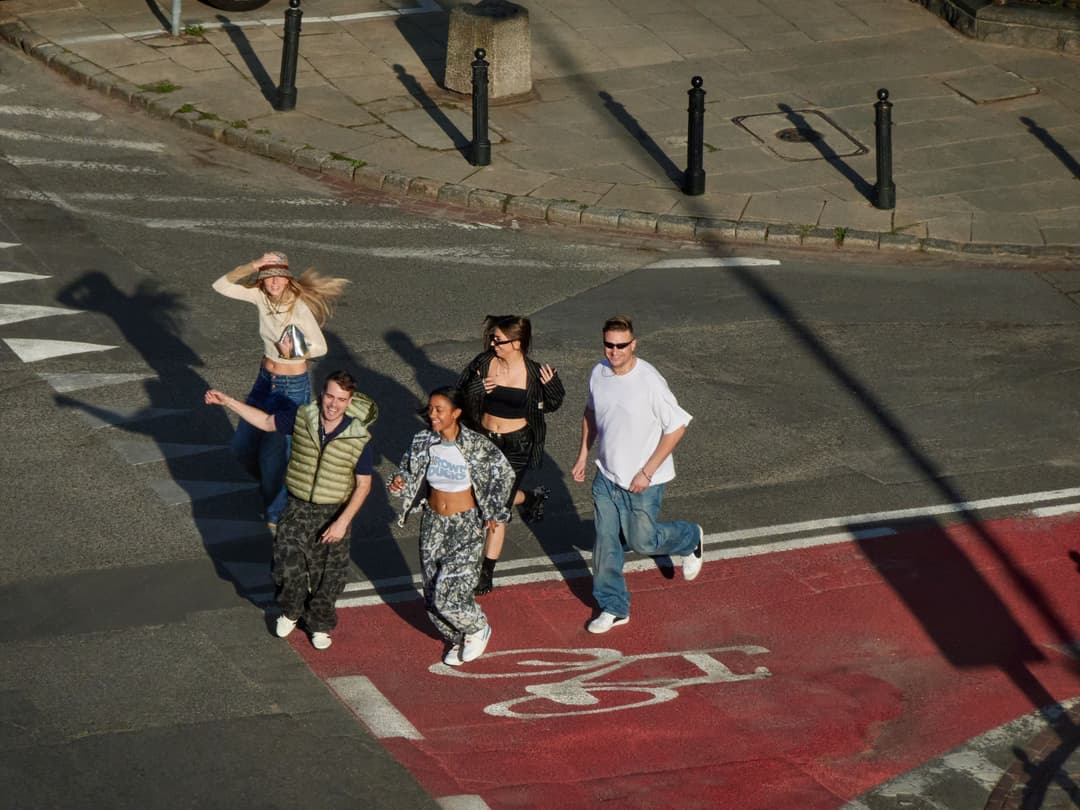 Cinq jeunes en tenue streetwear courent sur une place urbaine pavée aux marquages rouges et blancs, photographiés d'en haut par beau temps