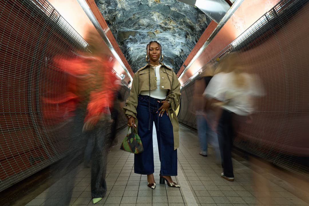 Femme en trench-coat beige et jean bleu foncé posant dans un passage souterrain urbain avec briques rouges et passants flous