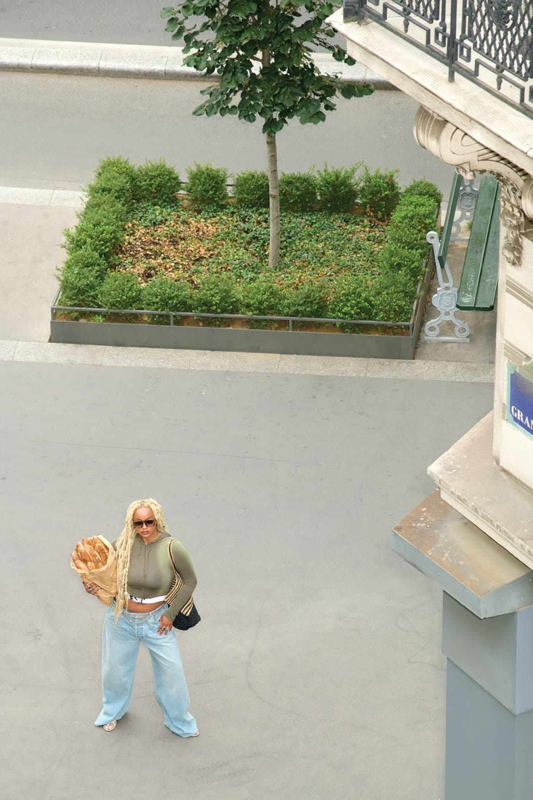 Femme en tenue streetwear posant dans une rue pavée avec baguette de pain, portant un crop top kaki, jeans clair et sac noir