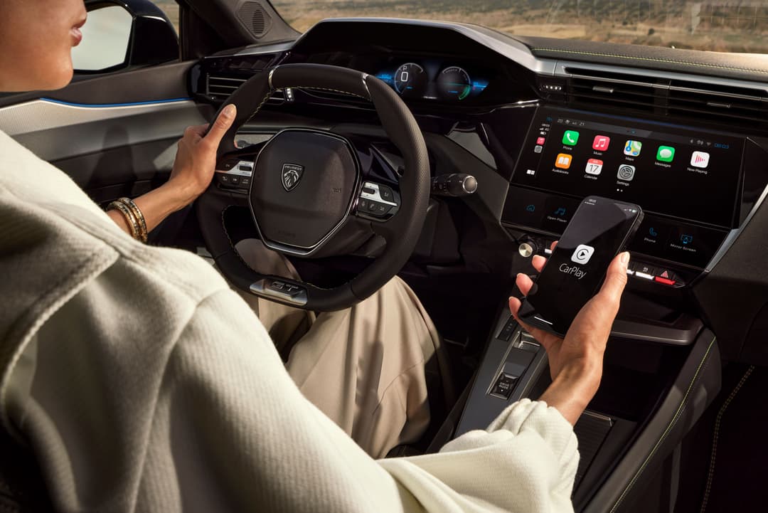 Intérieur de la Peugeot E-408 avec conductrice tenant le volant et un smartphone affichant Apple CarPlay sur l'écran tactile