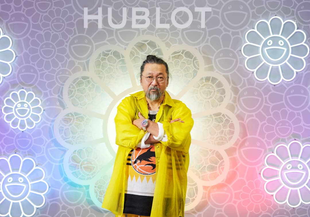 Takashi Murakami en manteau jaune vif devant un fond pastel orné de fleurs souriantes néon et de motifs circulaires