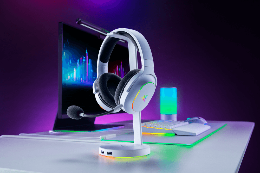 Casque gaming Razer Barracuda X Chroma blanc avec éclairages RGB multicolores, posé sur son support, entouré d'accessoires de gaming dans une ambiance violette et bleue