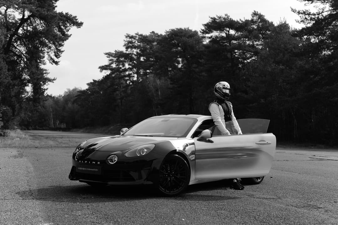Alpine A110 S monochromatique en noir et blanc, portière ouverte, avec un pilote en tenue de course devant une forêt