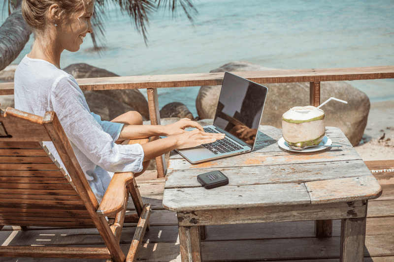Femme travaillant sur un laptop à une table en bois face à la mer, avec un routeur 5G NETGEAR Nighthawk M3 noir posé à proximité