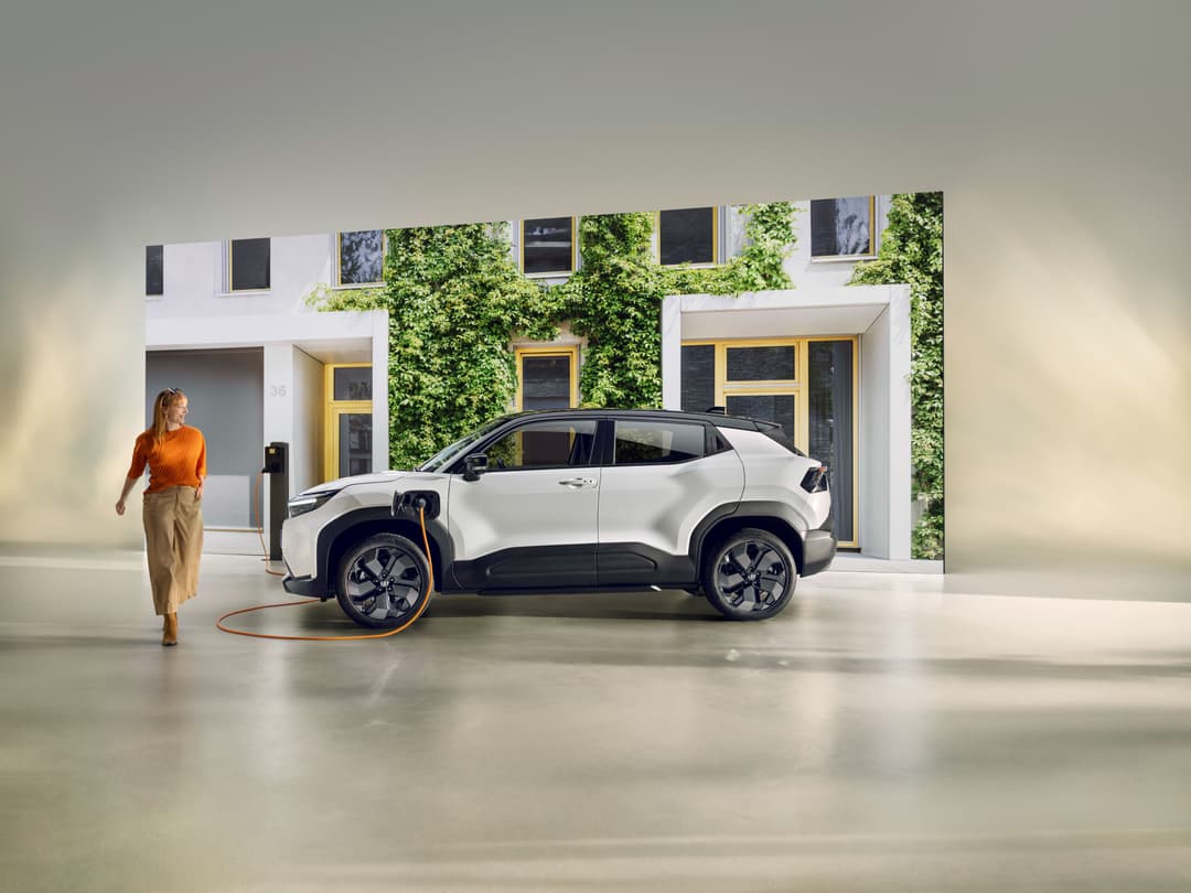 Toyota Urban Cruiser blanc et noir en charge devant un bâtiment moderne, une femme en pull orange à proximité
