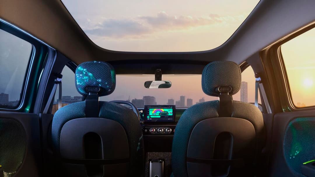 Intérieur de la Renault Twingo E-Tech electric prototype avec sièges aux motifs colorés bleu-turquoise, plancher en liège et toit panoramique ouvert sur un coucher de soleil urbain