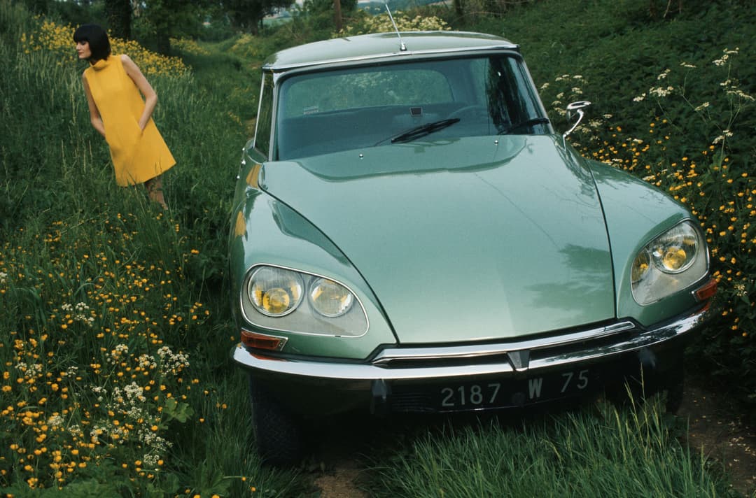 Citroën DS verte des années 1970 garée dans un champ de fleurs jaunes, avec une femme en robe jaune à proximité