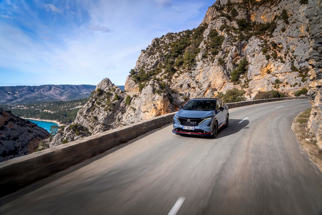 Nissan ARIYA NISMO blanche en mouvement sur une route sinueuse encadrée de falaises rocheuses avec un lac turquoise visible en arrière-plan