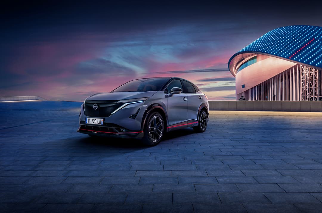 Nissan ARIYA NISMO grise avec liseré rouge, vue de profil au coucher de soleil face à un bâtiment moderne