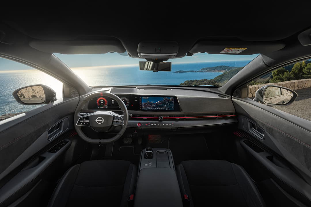 Habitacle du Nissan ARIYA NISMO avec sièges sportifs noirs, volant et écran tactile, vue sur la mer et les côtes depuis le pare-brise