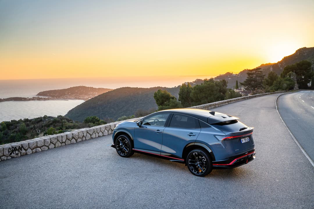 Nissan ARIYA NISMO bleu avec détails rouges, stationnée sur une route côtière sinueuse au coucher du soleil