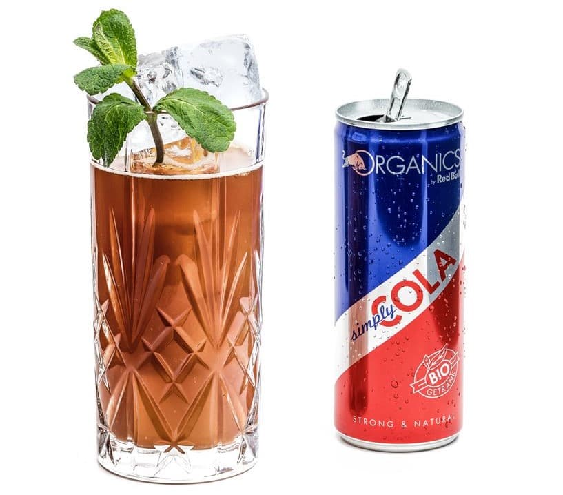 Verre de cocktail brun avec glaçons et feuilles de menthe à côté d'une canette ORGANICS by Red Bull Simply Cola bleu et rouge