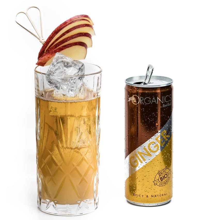 Verre highball rempli d'un cocktail jaune avec glaçons et décoration de lamelles de pomme rouge, à côté d'une canette ORGANICS Ginger by Red Bull
