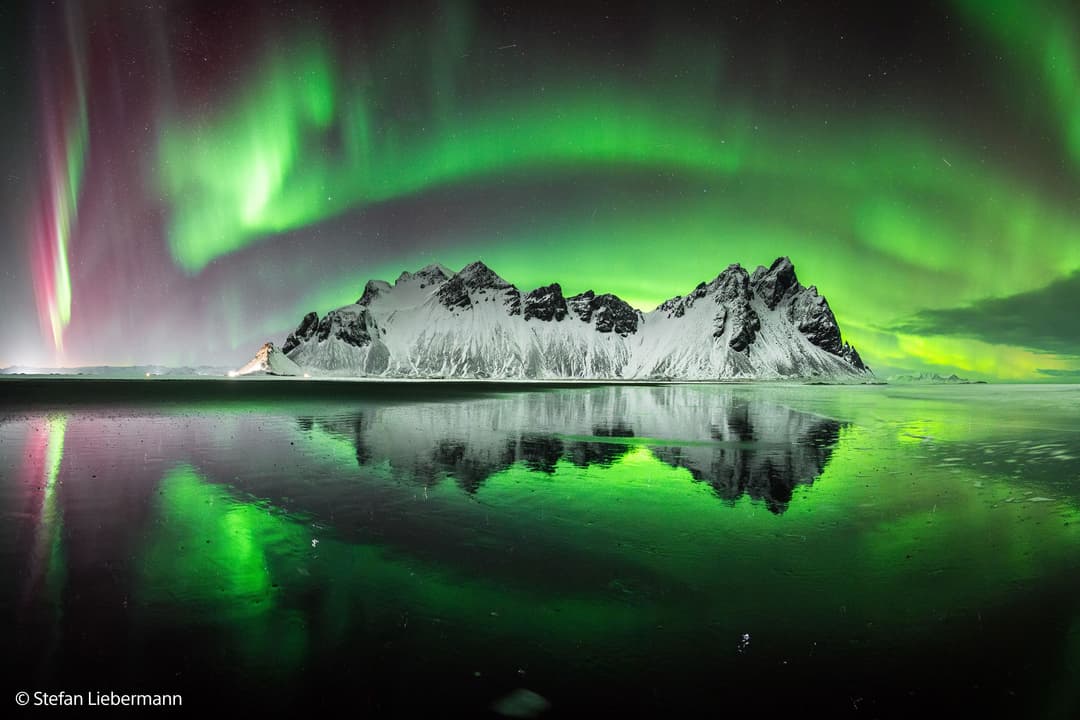 Montagne enneigée se reflétant dans une baie glacée sous aurore boréale verte et rose