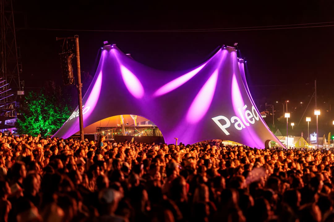 Foule nombreuse devant la scène du Paléo illuminée en violet et magenta, logo du festival visible sur la structure gonflable