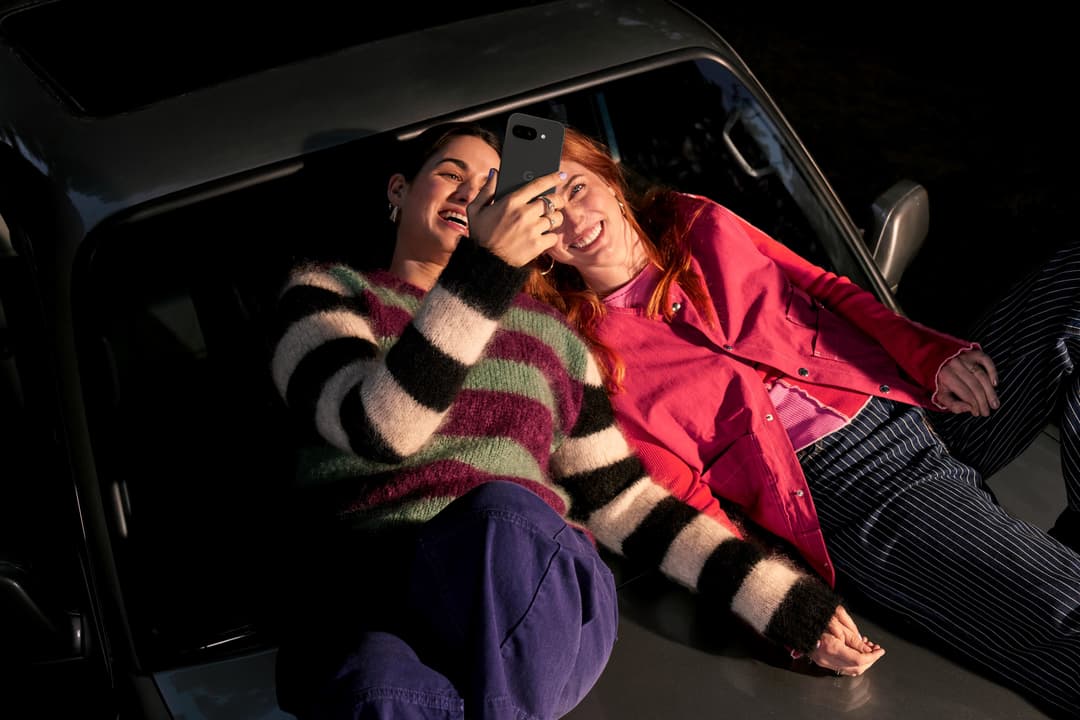 Deux femmes souriantes prennent un selfie dans une voiture avec le Pixel 9a, l'une en pull multicolore et l'autre en veste rouge