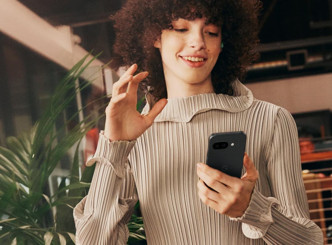 Personne tenant un Pixel 9a noir, souriante, dans un intérieur lumineux avec une plante verte