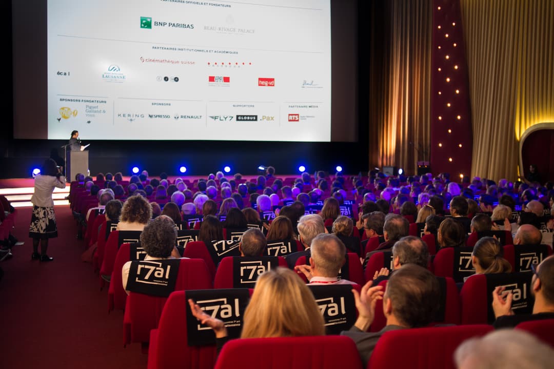 Salle de cinéma avec public assis sur des fauteuils rouges marqués du logo 7al, écran affichant les partenaires des Rencontres 7e Art, oratrice à la tribune, lumières bleues et rouges