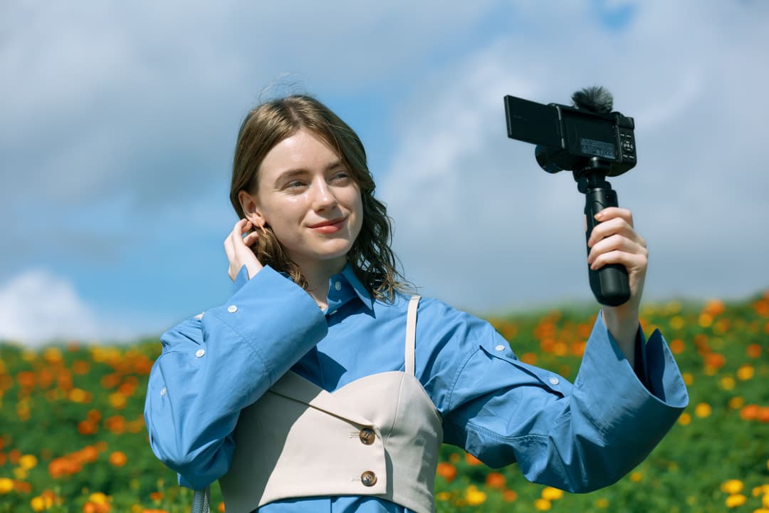 Jeune femme en chemise bleue tenant un caméscope Canon avec gimbal en extérieur dans un champ de fleurs