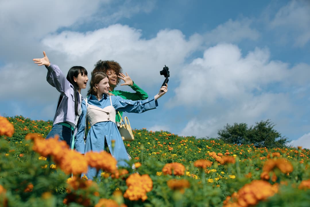 Trois jeunes femmes prenant un selfie dans un champ de fleurs orange sous un ciel bleu avec des nuages blancs