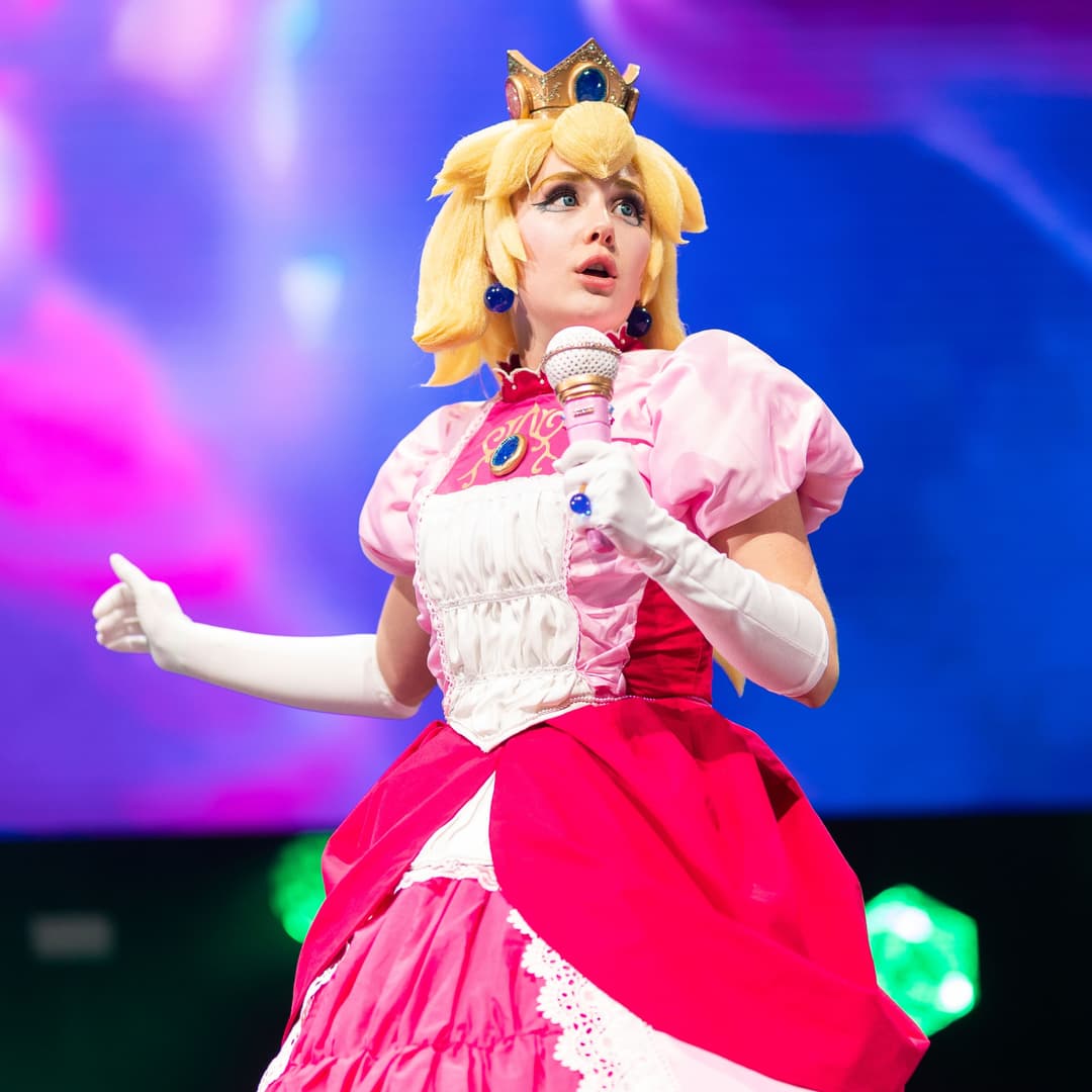 Performeuse en costume de princesse rose et rouge avec couronne, chantant au microphone sur scène éclairée de bleu et rose